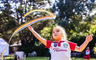A young Rainbow is stood outside, looking curious, as they swirl a ribbon above their head