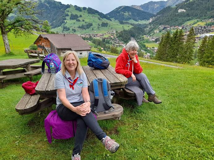 Two women smile at the camera while sitting on a bench on grass, surrounded by backpacks. In the background is a Swiss house, the village of Adelboden and green hills