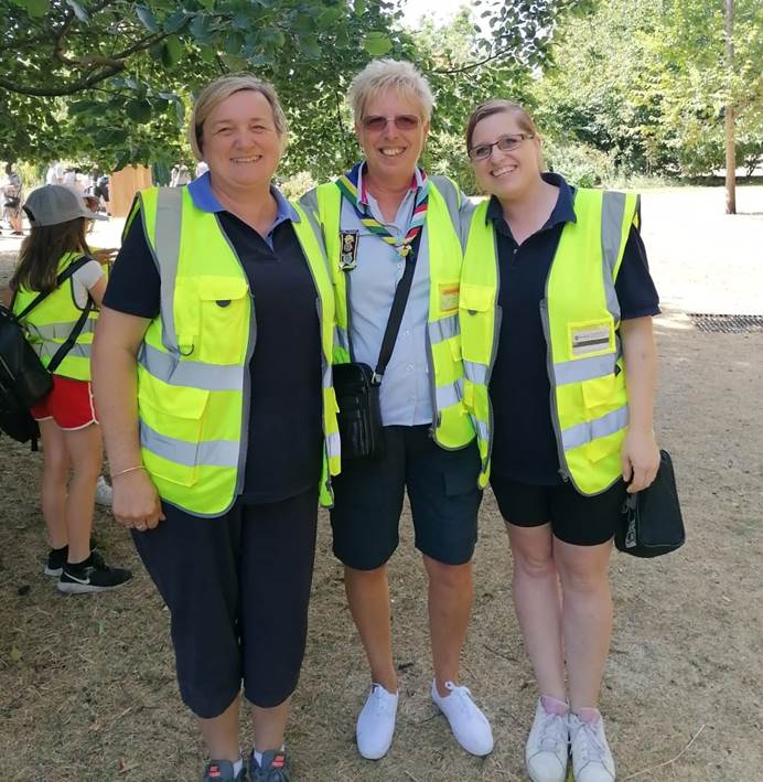 Three women wearing high vis vests stand smiling at the camera. It's sunny and they're in St James' Park, you can see trees, dry grass and a Brownie behind them