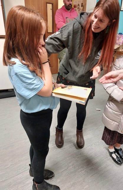 Chloë, who's white with mid length auburn hair, stands in a light blue top, black trousers with one hand in her mouth and the other holding a Brownie book, certificate and promise badge. Lynda, who's white and has long red hair, puts a hand on Chloë's shoulder and smiles at her. They're stood in a hall