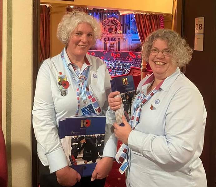 Two women stand in Girlguiding uniform, smiling at the camera. They're holding booklets for the festival. Behind them is the Royal Albert Hall's stage, with a big, lit up, poppy.