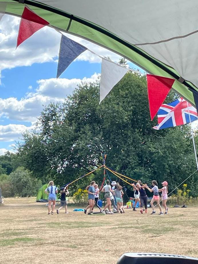 In the distance, Guides and Rangers are dancing around a maypole on dry grass. Behind them are trees. In the foreground is the edge of a tent and red, blue and white bunting