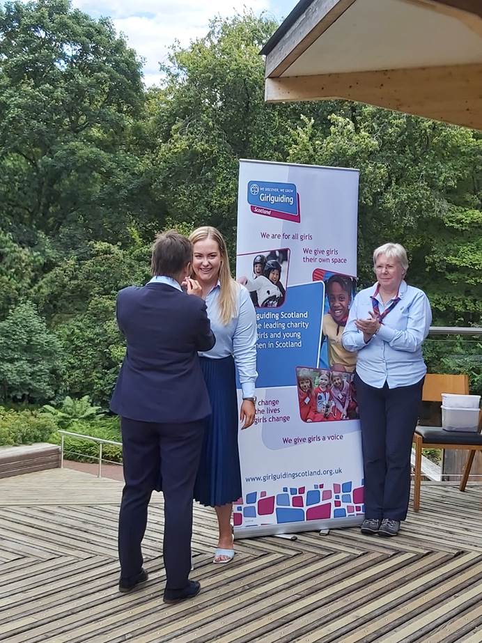 Emma, who is wearing a light blue shirt and long navy skirt, and has blonde hair, is smiling in front of a Girlguiding banner and facing towards the camera. Amanda, wearing a navy suit with her back to the camera, presents her with the award