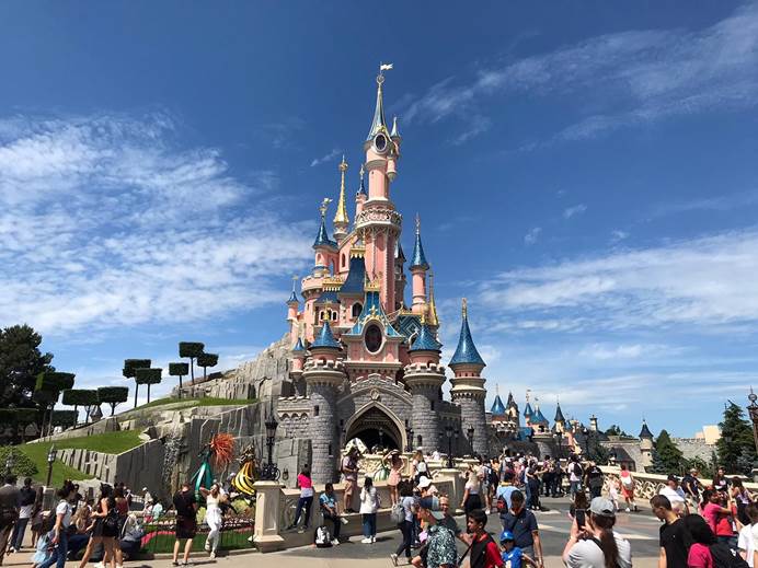 Lots of people are in the foreground, in front of Sleeping Beauty's castle. Behind the castle is blue sky, with a few white clouds