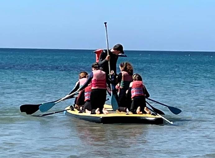 Four young members and an instructor, wear wetsuits and life jackets and face away from the camera on the mega paddleboard. They are paddling on a large expanse of water with blue sky above them