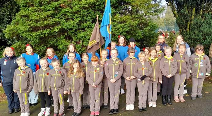 17 Brownies, 7 Guides and 3 leaders, all in uniform, stand in two lines looking at the camera. Two flags are being held upright in the middle of the photo. Behind them are trees