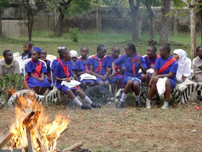 16 members of the Kenya Girl Guide's Association sit on painted tyres behind a campfire. Most of them are wearing blue dresses with red sashes and neckerchiefs. They're surrounded by grass and trees