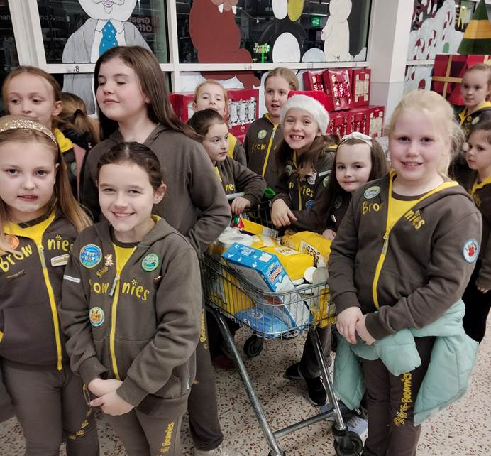 13 Brownies in uniform stand around a full shopping trolley in Asda. You can see the windows of the shop behind them. It's dark outside