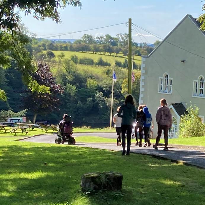 Kate, using a wheelchair, and a group of Guides to the right of her, move away from the camera at Waddow Hall. There are Peak District hills in the distance.