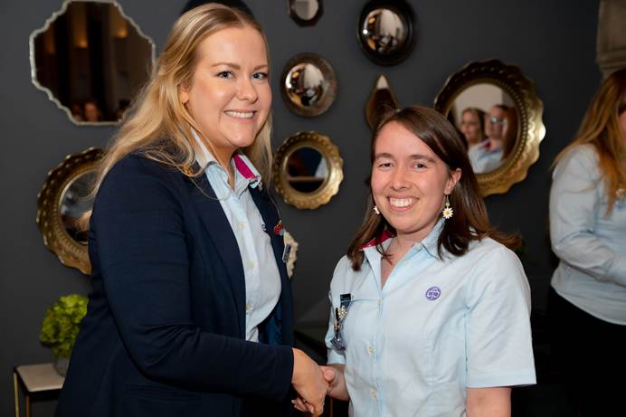 Katherine, stood on the right, receives her award from Emma Guthrie. They're both white, shaking hands, smiling at the camera and wearing uniform. They're inside