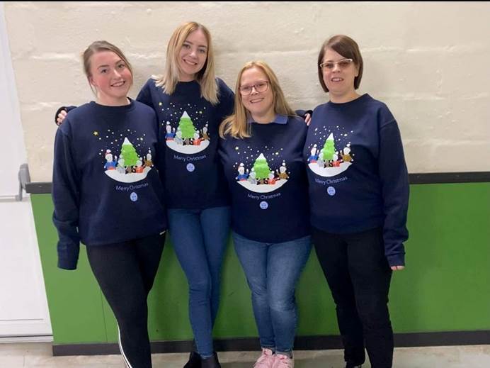 Four women stand in front of a wall, smiling at the camera. The wall is white in the top half, green in the bottom half. Their all wearing a Girlguiding Christmas jumper