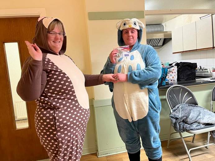 Two women wearing onesies are looking at the camera while doing a handshake and making the promise sign. Marion, on the right, holds a certificate and award. They're in a community hall and behind them is a door and a kitchen