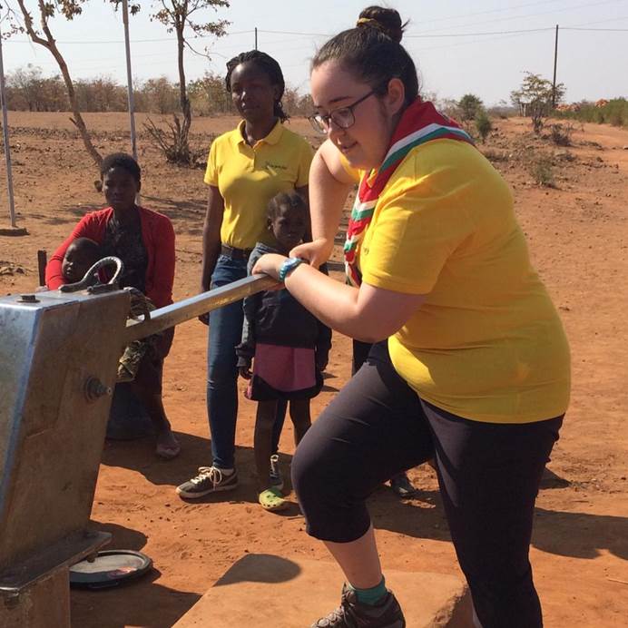 Nia is pumping water in a yellow t-shirt. She's white, has brown hair in a bun and glasses and is outside. 2 Black adults and 2 Black children watch her.  