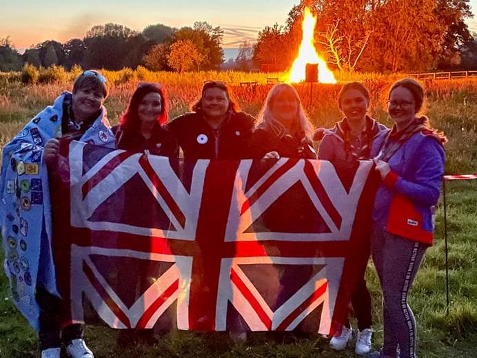 6 members look at the camera smiling, holding a Union Jack. In the background is a large campfire, making the whole photo orange